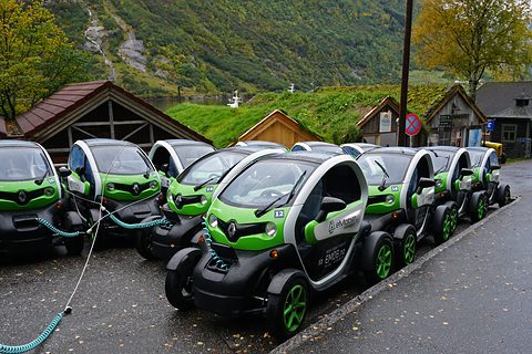 A row of green and white tandem two-seaters plugged in along a kerb below turf-roofed buildings and a steep hillside.