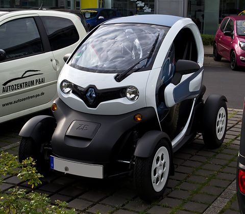 White and black tandem two-seater with blue trim parked on block paving among dealer cars. Opens a larger view