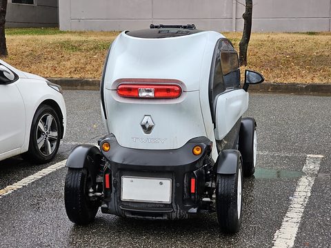 Rear of a white tandem two-seater with a red tail lamp and a blanked plate, parked on wet tarmac. Opens a larger view