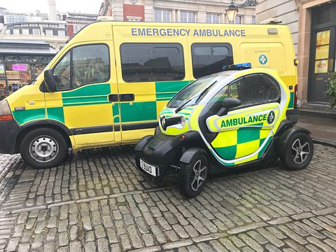 Yellow and green chequered tandem two-seater marked Ambulance, parked on cobbles next to a large ambulance.