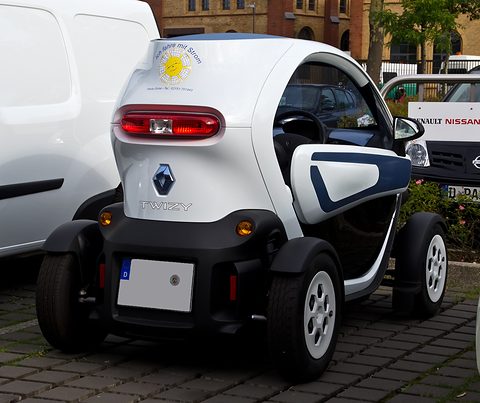 Rear of a white tandem two-seater with an ‘Ich fahre mit Strom’ sun sticker, parked between vans. Opens a larger view