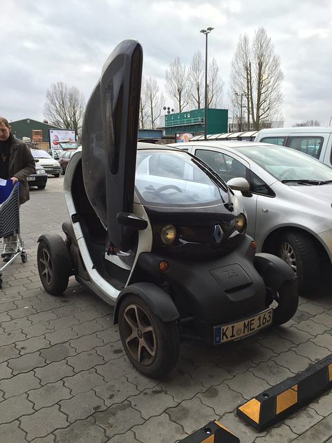 White and black tandem two-seater with its door raised upward, parked among cars under bare trees. Opens a larger view