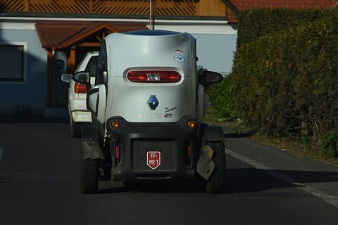 Rear of a grey tandem two-seater with a small plate, parked on a dark road by a hedge. Opens a larger view