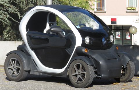 White and black tandem two-seater with dark wheels parked by a wall, trees and buildings behind. Opens a larger view