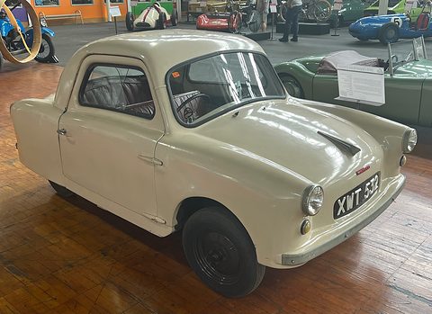 White rounded two-door microcar on a wooden museum floor, race cars in the background. Opens a larger view