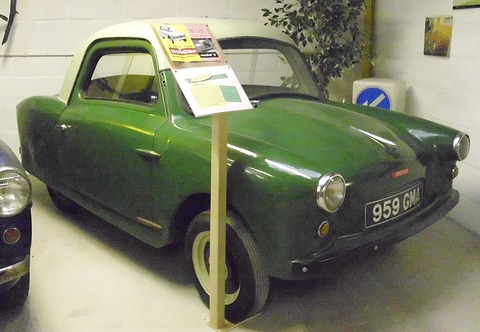Dark green microcar with cream roof, an information placard on a stick in front of it. Opens a larger view