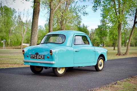 Back and side of a light-blue microcar with chrome bumper and small tail lamps on a park drive.