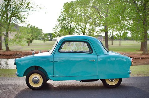 Light-blue rounded microcar seen side-on on a drive, pond and trees behind. Opens a larger view