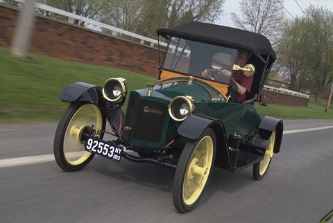 A green brass-era open car with yellow wheels and a black folding top driving along a country road, a driver at the wheel. Opens a larger view
