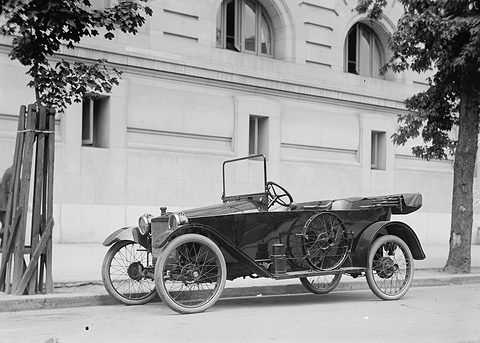 A small open tourer with wire wheels and a spare wheel on its side, parked at the kerb in front of an arched stone building. Opens a larger view