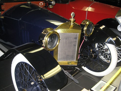 Close view of a brass radiator flanked by two large brass headlamps, black mudguards and white-walled wire wheels. Opens a larger view