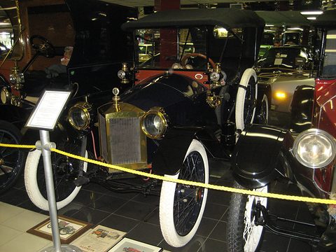 A black open tourer with brass radiator, brass lamps and white tyres behind a yellow rope, an information stand beside it. Opens a larger view