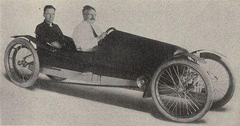 Two men in shirt sleeves sit one behind the other in a long, low, narrow car with wire wheels and cycle mudguards. Opens a larger view