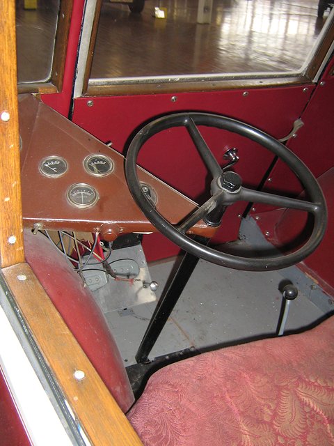 A black steering wheel on a long column, a small panel of round gauges and a red patterned bench seat seen through the door. Opens a larger view