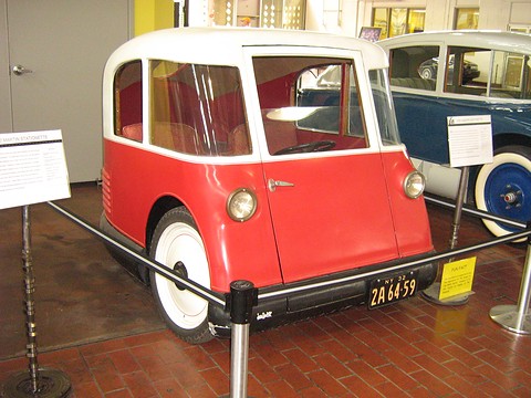 A tall red and cream three-wheeler with round headlamps and a single door, behind a barrier rope with placards. Opens a larger view