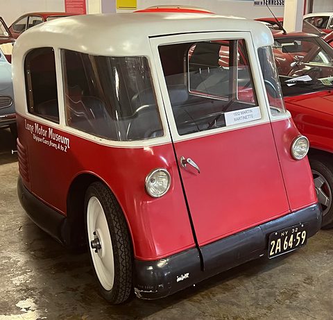 A tall rounded red and white three-wheeler with a single front door and a 1932 New York number plate. Opens a larger view