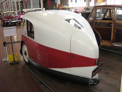 The rounded white and red tail of a three-wheeler with a number plate, a wood-bodied car beside it. Opens a larger view