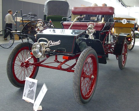 A small black open car with red wire wheels, brass lamps and a red button seat, a framed portrait propped at its front wheel. Opens a larger view