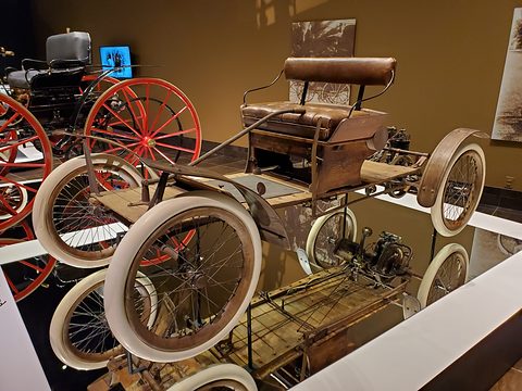 A bare wooden platform car on four white-tyred wire wheels, with a bench seat and a small engine at the back. Opens a larger view
