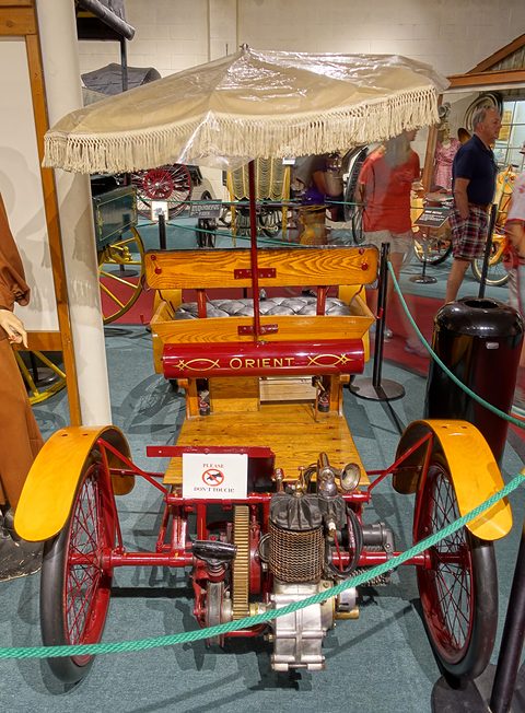 A wooden buckboard car with red wheels, yellow mudguards and a fringed parasol over its seat, roped off in a museum. Opens a larger view