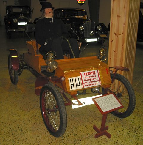 A wooden-bodied early car with a costumed mannequin at the tiller, a warning sign on its front and a placard beside it. Opens a larger view
