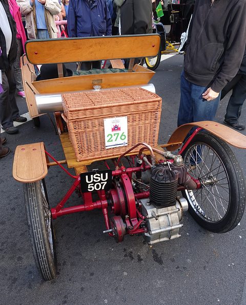Back view of a wooden buckboard car with an exposed single-cylinder engine, a wicker hamper and a black number plate. Opens a larger view