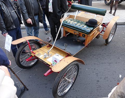 A varnished wooden buckboard car with a green buttoned bench seat and a bowler hat on it, spectators standing around. Opens a larger view