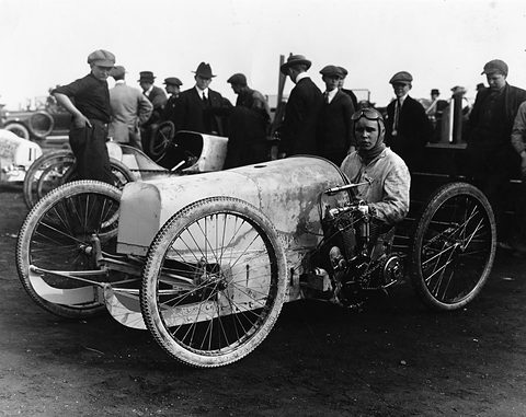 A goggled young man sits in a battered white racing cyclecar with wire wheels, men in caps and coats behind him. Opens a larger view