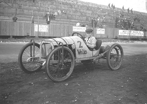 A young driver in a cap at the wheel of a small white racing car numbered 2, a wooden grandstand behind. Opens a larger view