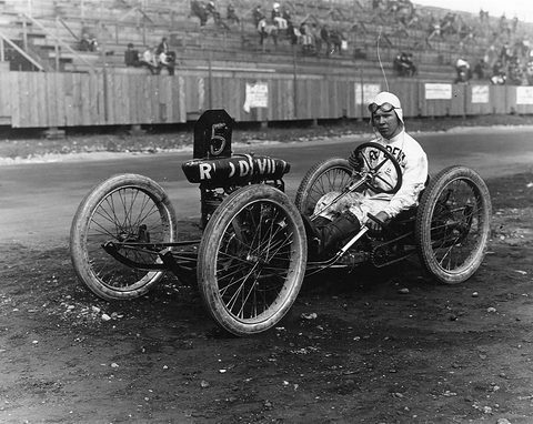 A driver in white overalls and helmet in a skeletal racing cyclecar with a number 5 board behind his seat. Opens a larger view