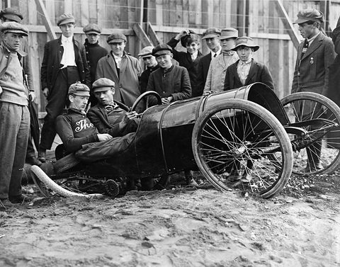A small barrel-bodied racing car lies tipped on the dirt, two young men in it, a crowd of men in caps standing behind. Opens a larger view