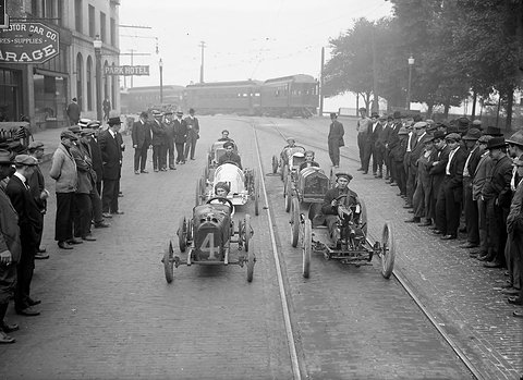 Two rows of small racing cars on a cobbled street with tram lines, crowds of men in caps lining both sides.