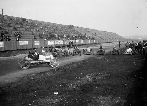 Small racing cars on a dirt track, one in the foreground, a long wooden grandstand behind. Opens a larger view
