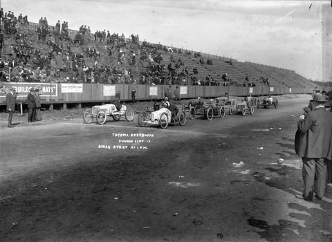 A row of small racing cars on a dirt track before a wooden grandstand full of spectators, men in coats in the foreground. Opens a larger view