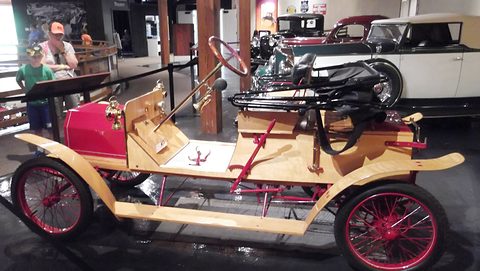 A light wooden open car with red wire wheels and a tall steering column, visitors and other cars behind it. Opens a larger view