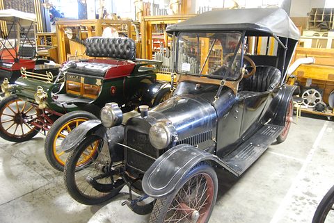 A black narrow-track open tourer with its top up, brass-trimmed early cars parked alongside on a concrete floor. Opens a larger view