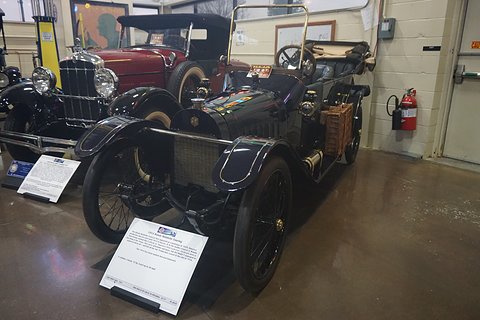 A small black open tourer with a wicker basket on its side and a placard on a stand in front, another car behind. Opens a larger view