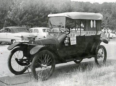 Black-and-white photograph of a narrow black tourer with a pale folding top and a show card on its windscreen, on grass. Opens a larger view