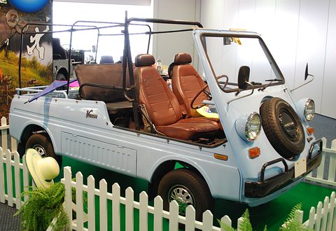 Pale blue doorless small utility with tan bucket seats, a nose-mounted spare wheel and bare top bows, behind a white picket fence.