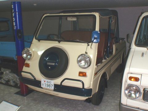 Front of a cream small utility with a spare wheel between round headlamps and a black canvas top, other cars on either side. Opens a larger view