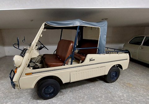 Cream doorless small utility with brown seats, a pale blue canvas top and a Vamos script on its flank, on a museum floor. Opens a larger view