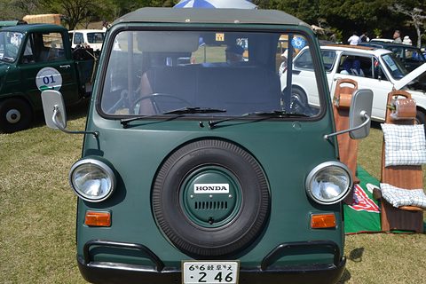 Head-on view of a dark green small utility with a spare wheel on its nose between round headlamps and a full canvas top, on grass. Opens a larger view