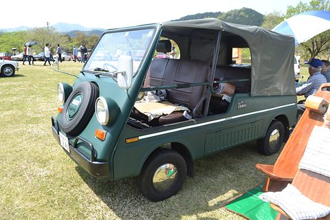 Dark green small utility with a full canvas top and a doorless side, parked on grass with deck chairs beside it and hills behind.