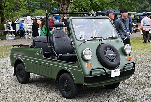 Olive green open small utility with a nose spare wheel, round headlamps and black seats, on gravel with visitors behind. Opens a larger view