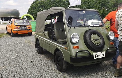 Olive green small utility with a canvas top and a nose spare wheel, on gravel, an orange Honda Z and visitors behind. Opens a larger view