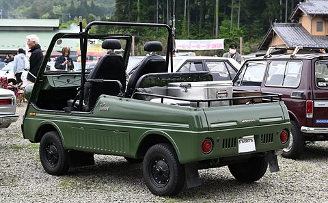 Rear quarter of an olive green open small utility with black bucket seats, roll hoops and a load bed, on gravel among parked cars. Opens a larger view