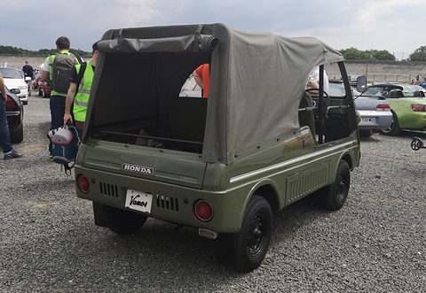 Rear quarter of an olive green small utility with a canvas top, HONDA lettering on its tailgate and rear guard rails, on gravel. Opens a larger view