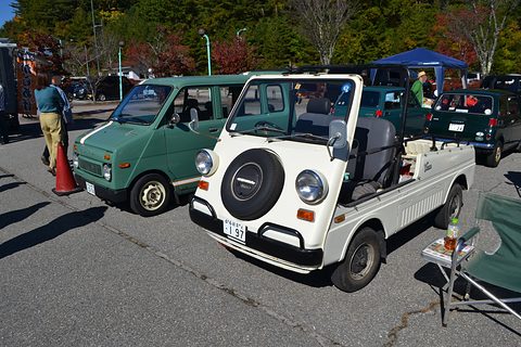 White doorless small utility with a nose-mounted spare wheel parked beside a green tall small van, visitors and tents around. Opens a larger view