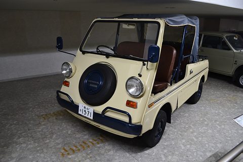 Cream small utility with a navy spare-wheel cover on its nose, round headlamps and blue guard rails, on a museum floor. Opens a larger view