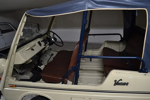 Doorless side of a cream small utility: brown front and rear bench seats, blue tubular guard rails and a simple dashboard under a navy top.